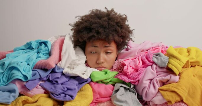 Stressed curly Afro American woman covered with heap of multicolored laundry feels tired after folding washed clothing has displeased expression brings house in order doesnt like mess in closet