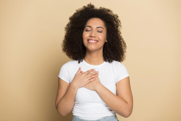 Happy millennial African American woman isolated on yellow background hold hands at heart chest feel thankful grateful. Smiling young biracial female feel religious superstitious. Faith concept.
