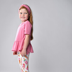 Friendly smiling little red-haired girl wearing summer fashion clothes, turning sideways looking at camera. Adorable little female model studio portrait shot. Positive emotions of children