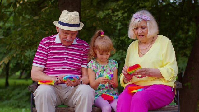 Smiling senior grandmother grandfather with granddaughter playing squeezing anti-stress silicone toy simple dimple game in park. Family with pop it bubble squishy game with buttons. Stress reliever