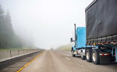 Big rigs semi trucks with semi trailers running on the wide one way highway road disappearing into the fog