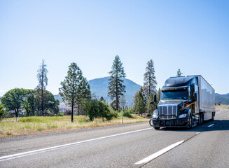 Black stylish big rig bonnet industrial semi truck with dry van semi trailer driving on the highway road with trees and mountain on background