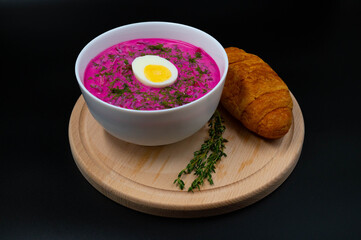 Summer cold beetroot soup (choldnik) with boiled egg served in a bowl on a wooden board with a croissant. Isolated black background.