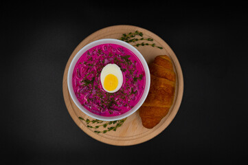 Summer cold beetroot soup (choldnik) with boiled egg served in a bowl on a wooden board with a croissant. Isolated black background. Top view.