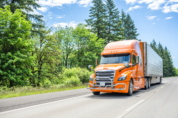 Fototapeta na wymiar Catchy orange big rig semi truck with pipe grille guard transporting frozen cargo in refrigerated semi trailer driving on the highway road with green forest on the side