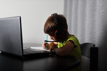 Serious boy in shadow listening to lessons on laptop and writing in notepad with a pen at home. Online education and back to school concept