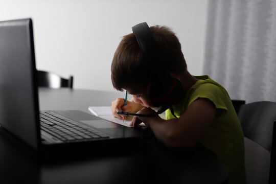 Boy sitting in shadow on laptop and wears headphones on ears and writing in notebook with pen. Online education and back to school concept - Powered by Adobe