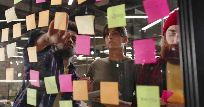 Creative multiethnic team of developers brainstorming ideas, putting stickers on glass board. Students working on project 4k footage
