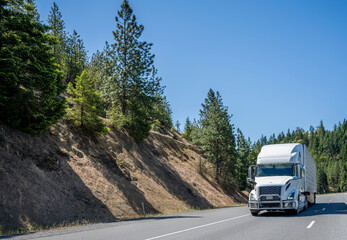 Industrial grade professional commercial big rig white semi truck transporting cargo in refrigerated semi trailer driving on the mountain road with trees on the hillside
