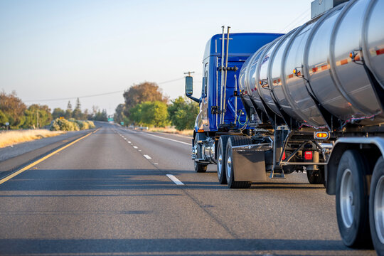 Low profile cab roof blue big rig long haul semi truck transporting liquid cargo in armored tank semi trailer driving on the one way divided highway road