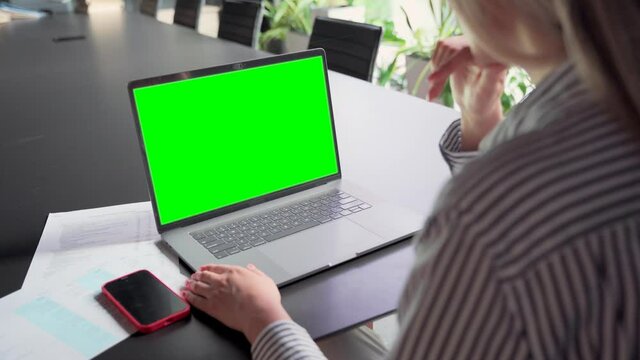 Young Caucasian businesswoman ceo executive manager using typing email letter using laptop computer with empty blank green mockup screen for advertising at desk. Business concept. Over shoulder view.