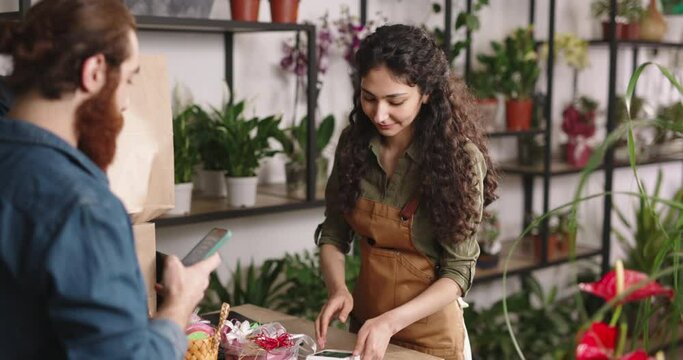 Customer paying for purchase in flower shop using nfc mobile technology. Asian female entrepreneur positively smiling - small business 4k footage