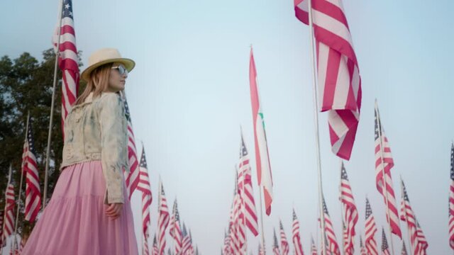Cinematic slow motion of young stylish woman walking between many American flags with blue sky background. Low angel side view of model looking at the national symbol of United States of America, 4K
