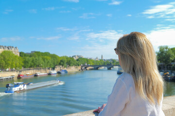 Fototapeta na wymiar Blonde woman from behind on a bridge in Paris looking at the Seine river in summer. Barge blurred deliberately in the background.
