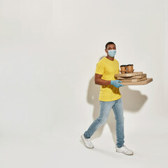 Full length shot of delivery man wearing protective mask and gloves looking at camera, holding many pizza boxes and drinks isolated over gray background