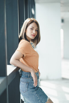 Stylish Asian girl wearing orange t-shirt and jean skirt posing in empty room, urban clothing style. Street photography.