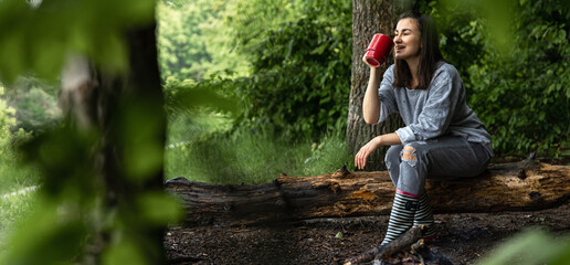 Naklejka na ściany i meble A girl with a cup of hot drink in the forest is resting near the fire.