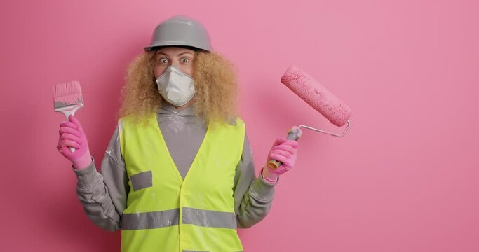 Pleased curly haired female painter jumps from joy holds painting tools ready for apartment remodeling wears hardhat uniform looks away on blank empty space against pink background. Repair concept