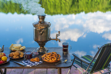 Obraz na płótnie Canvas Vintage metal tea samovar with white smoke and food on the table near the calm water lake in green forest at morning