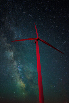 Astrophotography shot of a wind turbine and the Milky Way Galaxy as a shooting star passes behind the red wind turbine