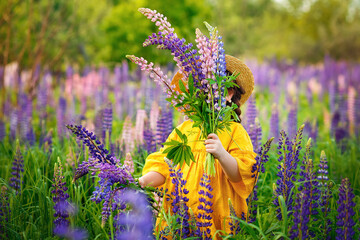 A child in a hat with a bouquet of lupin flowers. A girl in a yellow dress, a straw hat, hides behind a bouquet of lilac flowers in a flower meadow. Field of lupins.