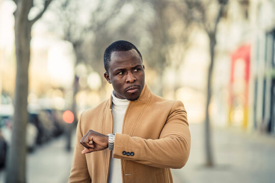 Serious black man checking time on wristwatch while strolling on sidewalk