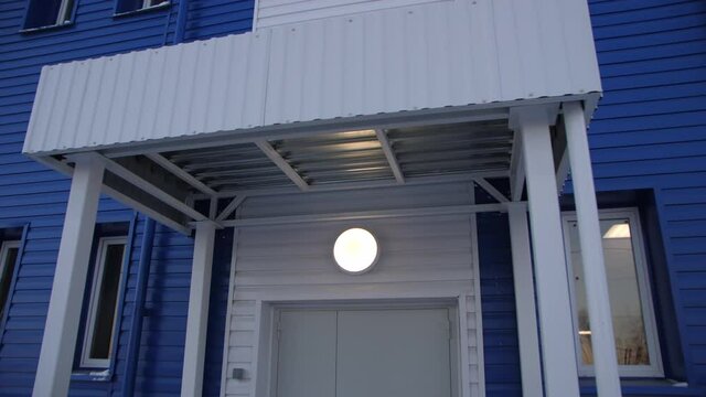 Closed entrance to contemporary blue workshop building with powerful glowing lamp above door in winter evening zoom in