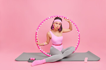 Slim fitness woman has training with hula hoop dressed in sportswear engaged in sport poses on mat looks attentively at camera isolated over pink background. Active lifestyle and home workout