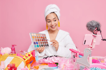 Beautiful pleased Asian woman holds eye shadow palette going to do make up talks with followers wears white bathrobe towel over head focused at smartphone webcam records cosmetic product review