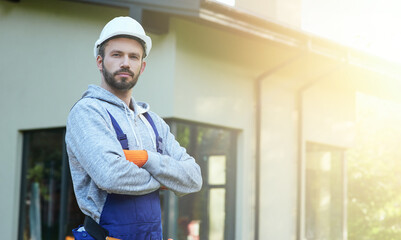 Portrait of young bearded male engineer in hard hat looking at camera, posing outdoors while working on cottage construction