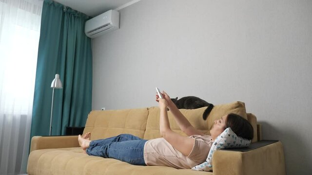 Woman turns on the air conditioner by the remote control, lying on the couch with a cat.