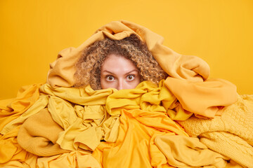 Horizontal shot of surprised curly haired woman covered with various clothing items cleans out...