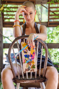Playful portrait of a young gorgeous female painter artist looking and smiling at camera, sitting on an old wooden chair, holding palette and a paintbrush.