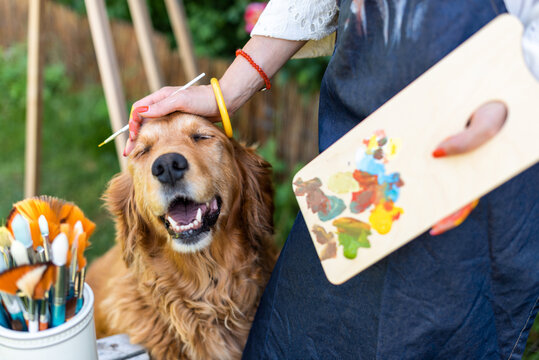 Young female artist working on her art canvas painting outdoors in her garden with golden retriever keeping her company. Creative hobbies concept.