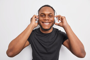 Cheerful handsome man with dark skin smiles broadly enjoys listening music via wireless headphones...