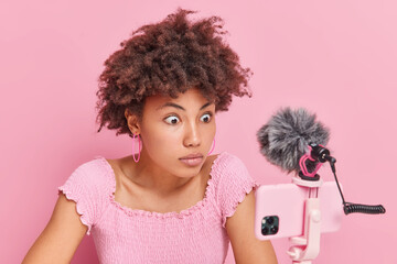 Shocked Afro American woman with curly hair stares at phone webcam records live stream video has...