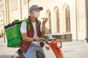 Contactless delivery during quarantine. Young asian male courier with thermo bag wearing face protective mask talking by mobile phone