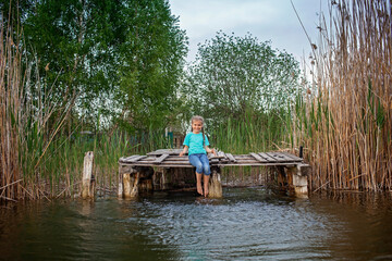 Cute preteen girl sits on a wooden pier by the lake and stirs feet in the water, happy childhood and summertime, escape to nature, relax without gadget, outdoor lifestyle