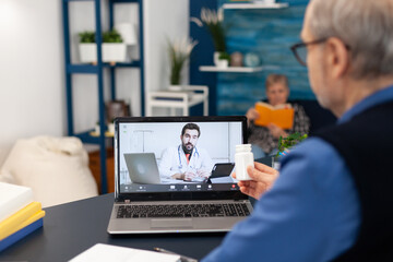 Young doctor talking about treatment with senior man during virtual examination. Elderly man discussing with healthcare practitioner in the course of remote call and wife is reading a book on sofa.