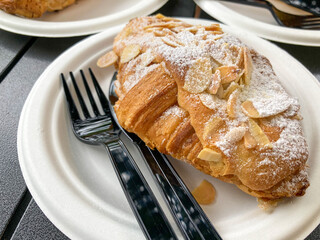 Croissant with icy sugar and sliced almond nut as topping put on white paper plate and black plastic fork and knife in coffee cafe