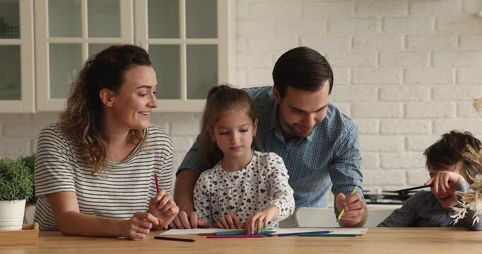 Caring young couple parents having fun with joyful cute small son and daughter, enjoying drawing in paper sketchbook together in kitchen, happy two generations family creative domestic pastime.