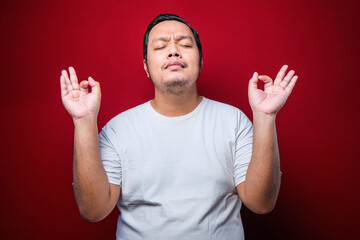 Fototapeta premium Young asian man wearing white t-shirt relax and smiling with eyes closed doing meditation gesture with fingers. Yoga concept.