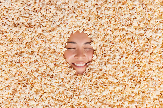 Positive young Asian woman buried in tasty popcorn smiles gladfully shows white teeth feels cheerful while watches comedy film. Food background. Creative image. Cinema and healthy snack concept