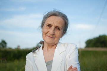 Portrait of senior woman with grey hair and face with wrinkles wearing white jacket and relaxing at park during sunny day