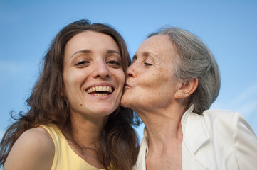 Senior mother with gray hair with her adult daughter looking at the camera in the garden and hugging each other during sunny day outdoors