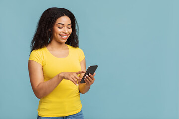 Cool app. Happy african american woman using smartphone, standing over blue studio background with free space