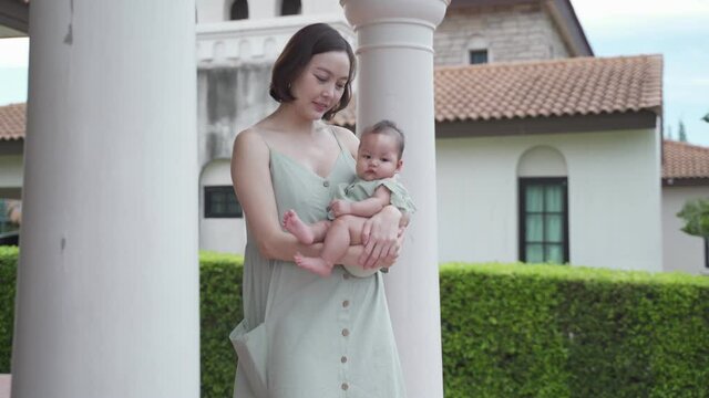 portrait of asian woman mother holding baby infant on balcony terrace at luxury home looking away the morning view. happy smile 30's female parent and 6 months baby at home near garden daytime.
