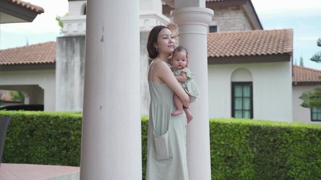 portrait of asian woman mother holding baby infant on balcony terrace at luxury home looking away the morning view. happy smile 30's female parent and 6 months baby at home near garden daytime. - Powered by Adobe