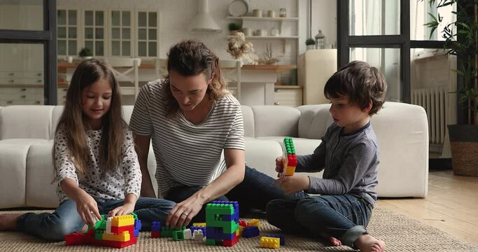 Caring young female babysitter or loving mother sitting on floor carpet with small children boy and girl, communicating enjoying playing toys together in modern living room, kids daycare concept.