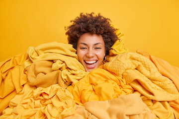 Positive cheerful curly haied young African American woman covered with heap of clothing after...